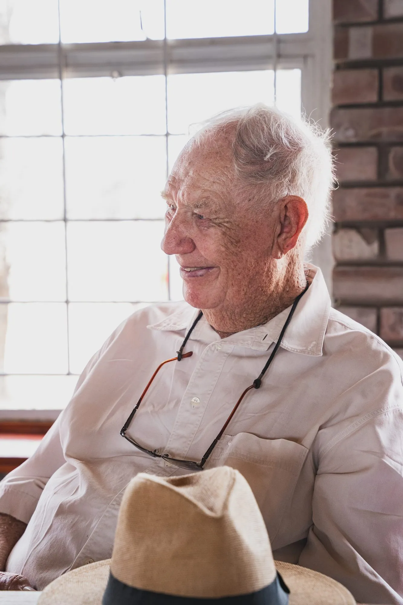 An elderly guest with his hat on his lap, lit by a window